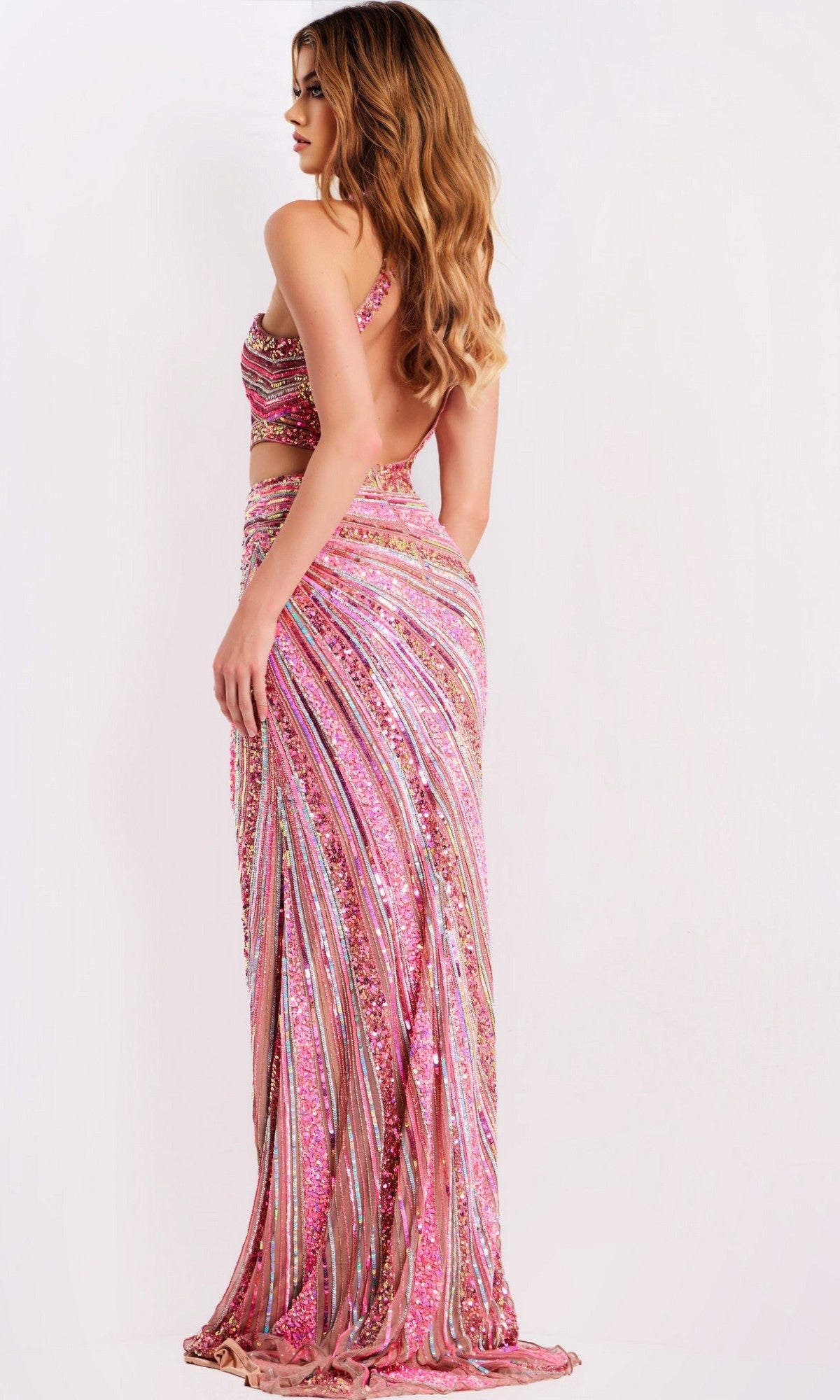 A woman in a pink, sequined Jovani 49266 long prom dress with colorful vertical and diagonal stripes stands sideways against a plain background.